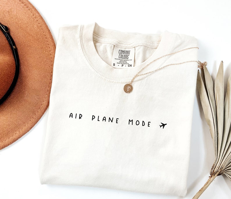 Peut inclure: Un t-shirt blanc avec le texte "AIR PLANE MODE" et une petite ic&ocirc;ne d'avion imprim&eacute;e sur le devant. Le t-shirt est pli&eacute; et pos&eacute; &agrave; plat sur une surface blanche.