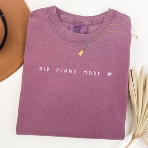 Peut inclure: Un t-shirt mauve avec le texte "AIR PLANE MODE" et un petit avion imprim&eacute; en blanc sur le devant.