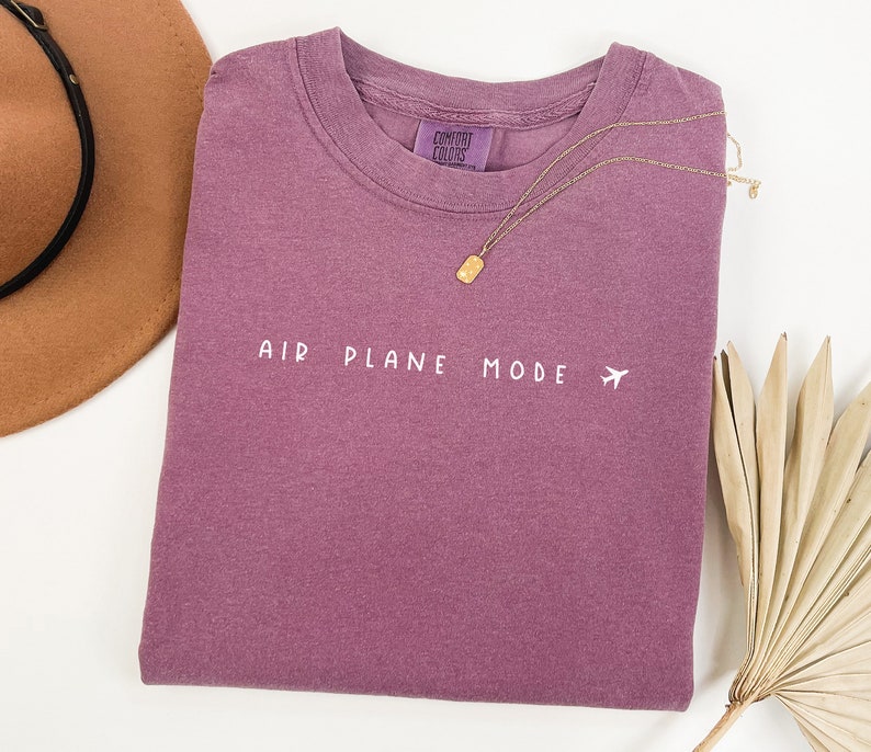 Peut inclure: Un t-shirt mauve avec le texte "AIR PLANE MODE" et un petit avion imprim&eacute; en blanc sur le devant.