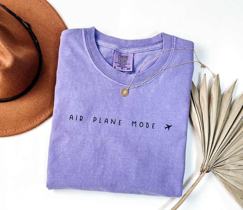 Peut inclure: Un t-shirt de couleur lavande avec le texte "AIR PLANE MODE" et un petit avion graphique. Le t-shirt est pli&eacute; et pos&eacute; &agrave; plat sur une surface blanche.