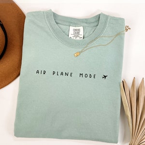 Peut inclure: Un t-shirt bleu clair avec le texte "AIR PLANE MODE" et une petite ic&ocirc;ne d'avion imprim&eacute;e en noir.