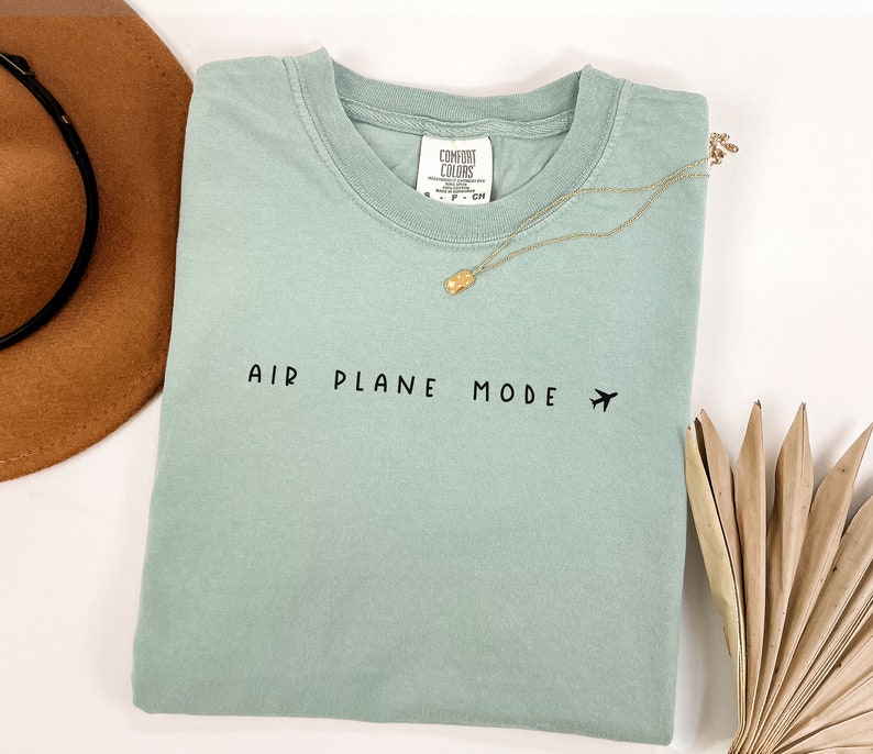 Peut inclure: Un t-shirt bleu clair avec le texte "AIR PLANE MODE" et une petite ic&ocirc;ne d'avion imprim&eacute;e en noir.
