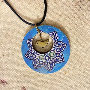 May include: A round blue pendant necklace with a floral design and a small gold-colored bird charm. The pendant has a black cord and a gold-colored clasp. The floral design features shades of blue, white, and green.