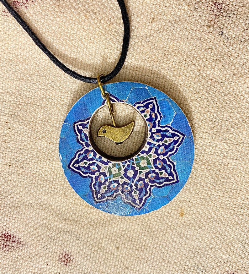 May include: A round blue pendant necklace with a floral design and a small gold-colored bird charm. The pendant has a black cord and a gold-colored clasp. The floral design features shades of blue, white, and green.