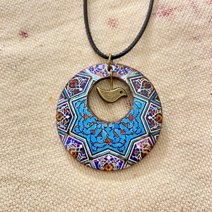 May include: A circular pendant necklace with a detailed, colorful mosaic design. The pendant features a central blue star pattern and a small, bronze-colored bird charm. The necklace has a black cord.