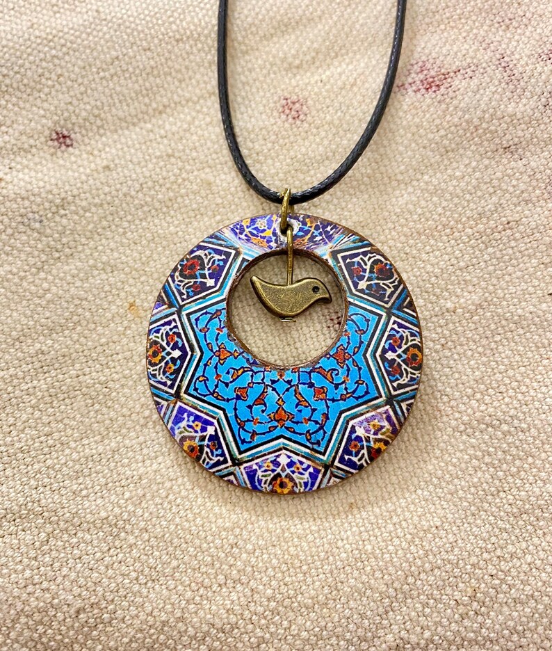 May include: A circular pendant necklace with a detailed, colorful mosaic design. The pendant features a central blue star pattern and a small, bronze-colored bird charm. The necklace has a black cord.