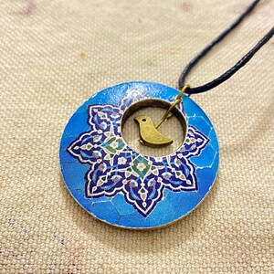 May include: A blue pendant necklace with a circular design. The pendant features a mosaic-style pattern in shades of blue, white, and green, with a small gold-colored bird charm in the center. The necklace has a black cord.