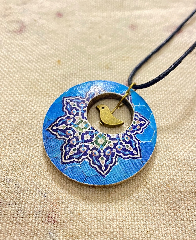 May include: A blue pendant necklace with a circular design. The pendant features a mosaic-style pattern in shades of blue, white, and green, with a small gold-colored bird charm in the center. The necklace has a black cord.