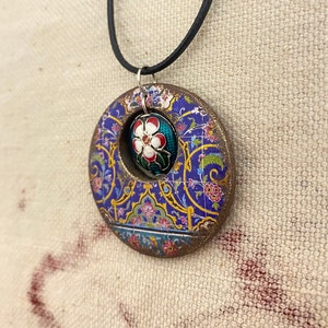 May include: A circular pendant necklace with a floral design. The pendant features a blue background with gold and pink floral patterns. A smaller, oval-shaped pendant with a white flower is set within the larger circle. The necklace has a black cord.