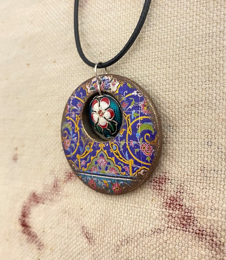 May include: A circular pendant necklace with a floral design. The pendant features a blue background with gold and pink floral patterns. A smaller, oval-shaped pendant with a white flower is set within the larger circle. The necklace has a black cord.
