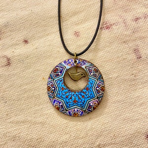 May include: A circular pendant necklace with a black cord. The pendant features a blue, white, and red geometric pattern with a small bronze bird charm in the center. The necklace is displayed on a beige fabric background.