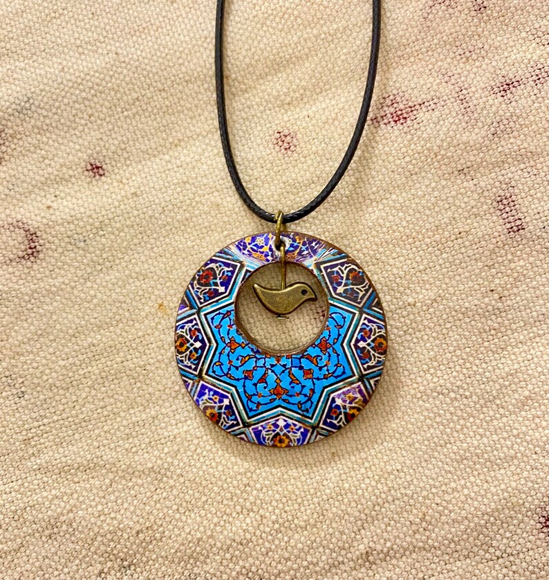 May include: A circular pendant necklace with a black cord. The pendant features a blue, white, and red geometric pattern with a small bronze bird charm in the center. The necklace is displayed on a beige fabric background.