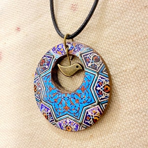 May include: A circular pendant necklace with a detailed mosaic pattern in blue, orange, and white. A small, bronze-colored bird charm hangs in the center. The pendant is suspended from a black cord.