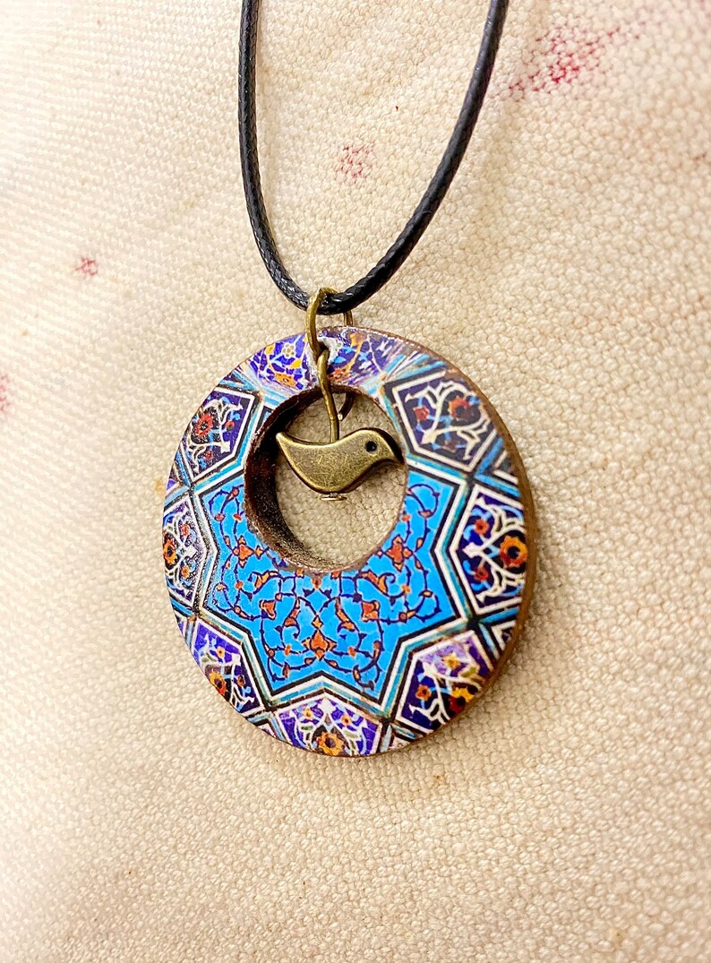 May include: A circular pendant necklace with a detailed mosaic pattern in blue, orange, and white. A small, bronze-colored bird charm hangs in the center. The pendant is suspended from a black cord.