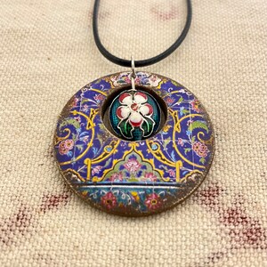May include: A round pendant necklace with a blue, gold, and floral design. The pendant has a central opening with a smaller, round, floral-patterned charm. The necklace has a black cord.