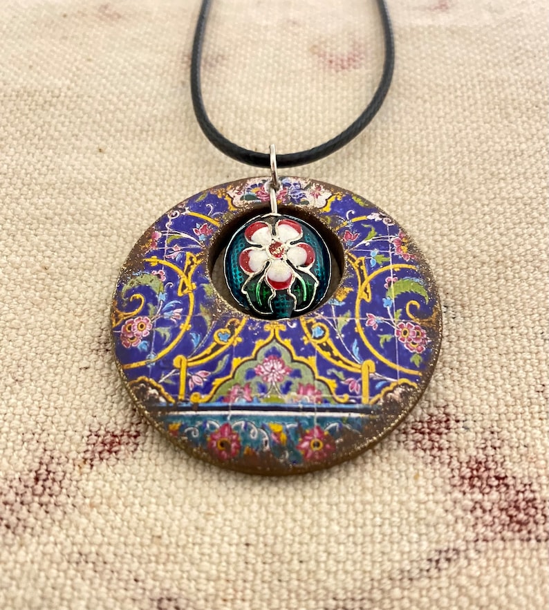 May include: A round pendant necklace with a blue, gold, and floral design. The pendant has a central opening with a smaller, round, floral-patterned charm. The necklace has a black cord.