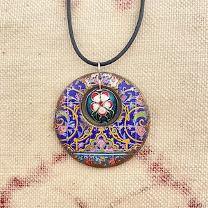 May include: A round pendant necklace with a blue floral design and a central flower motif. The pendant is suspended from a black cord. The pendant's design includes red, white, and yellow accents.