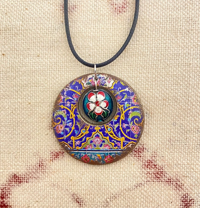 May include: A round pendant necklace with a blue floral design and a central flower motif. The pendant is suspended from a black cord. The pendant's design includes red, white, and yellow accents.