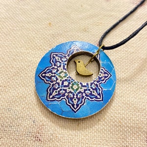 May include: A round pendant necklace with a blue and white floral design. A small gold-colored bird charm is centered within the pendant. The necklace has a black cord.