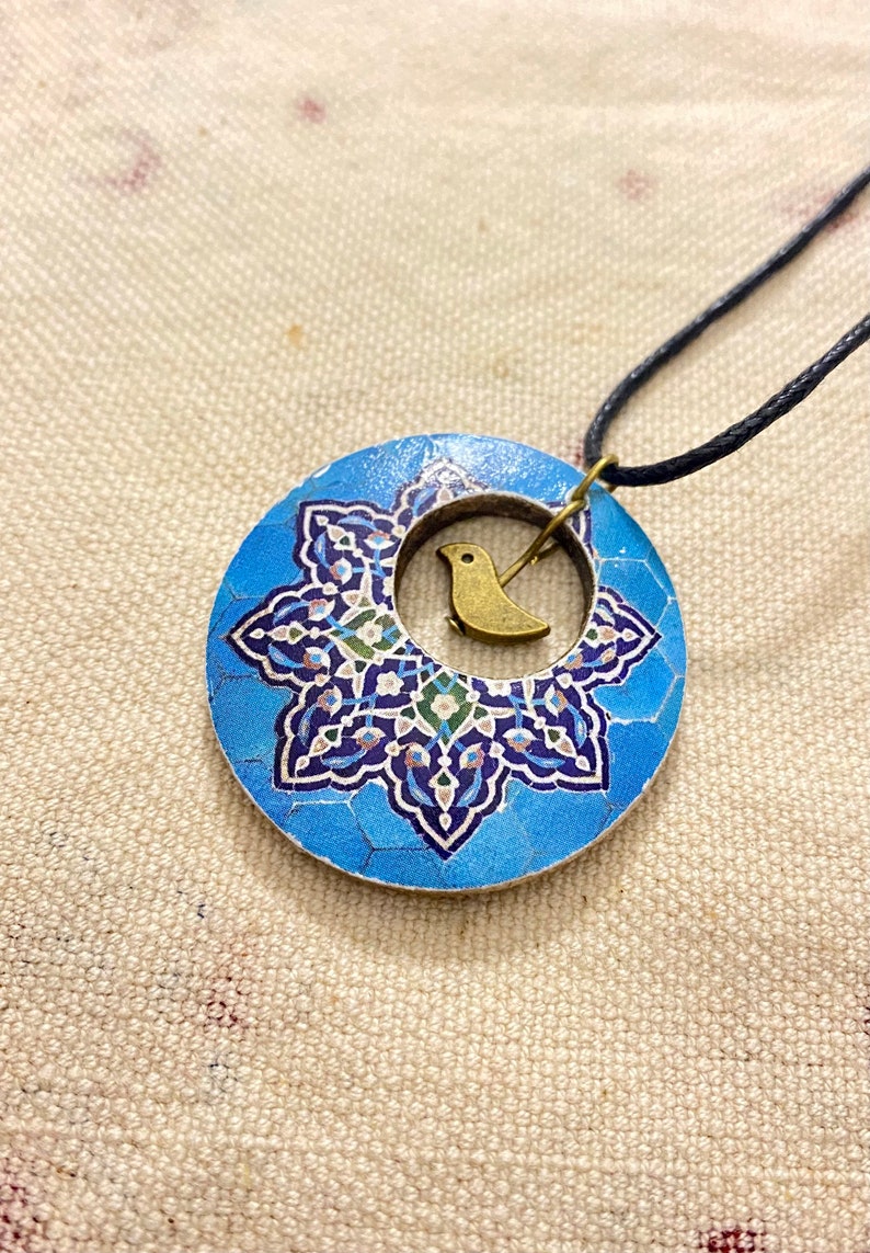 May include: A round pendant necklace with a blue and white floral design. A small gold-colored bird charm is centered within the pendant. The necklace has a black cord.