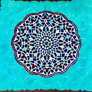 以下が含まれることがあります： 青、白、緑、オレンジの色合いで描かれた、中央に花の模様がある装飾的なタイル。タイルは、ターコイズ色のハニカム模様のボーダーで囲まれています。デザインは対称的で複雑であり、詳細な芸術的デザインです。サイズはセンチメートルです。