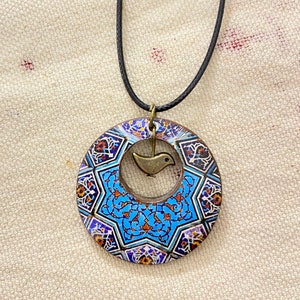 May include: A circular pendant necklace with a black cord. The pendant features a blue and white geometric design with a small bird charm in the center. The pendant has a bronze-colored metal frame.