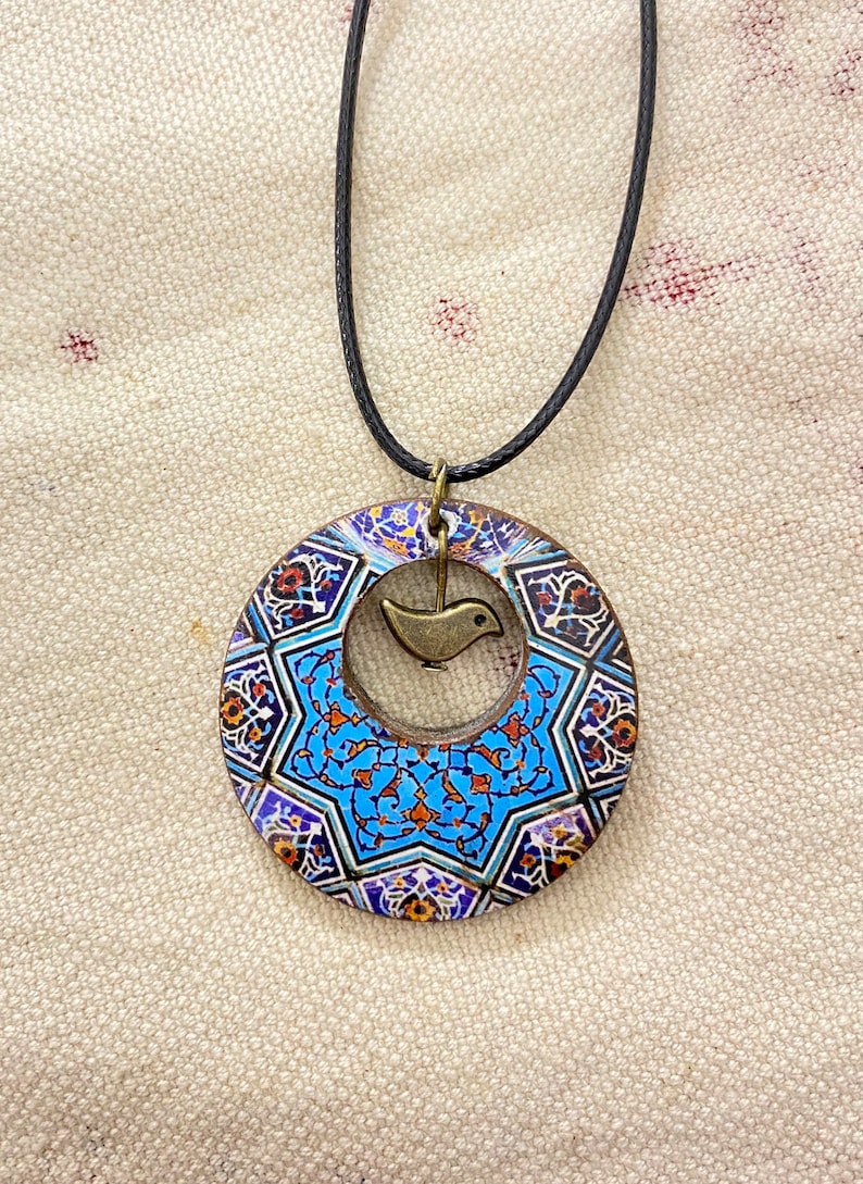 May include: A circular pendant necklace with a black cord. The pendant features a blue and white geometric design with a small bird charm in the center. The pendant has a bronze-colored metal frame.
