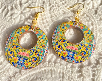 Puede incluir: Pendientes circulares con un vibrante patrón floral y geométrico en amarillo, azul y rojo. Los pendientes tienen ganchos dorados y un anillo interior marrón, creando un contraste llamativo. El diseño recuerda a los azulejos tradicionales.