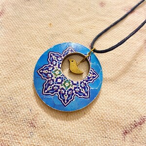 May include: A blue pendant necklace with a floral design and a small gold-colored bird charm. The pendant is round and has a dark blue cord. The necklace is a piece of jewelry.