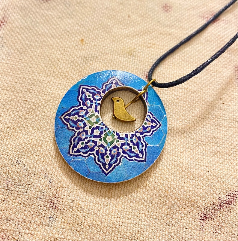 May include: A blue pendant necklace with a floral design and a small gold-colored bird charm. The pendant is round and has a dark blue cord. The necklace is a piece of jewelry.