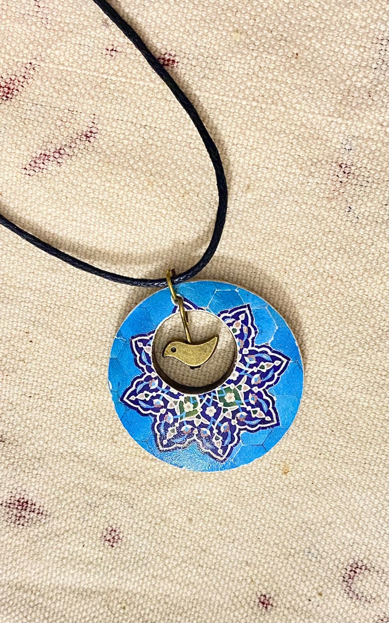 May include: A blue pendant necklace with a circular design featuring a floral pattern in shades of blue, white, and green. A small gold-colored bird charm hangs in the center. The necklace has a black cord.