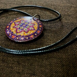 May include: A round pendant necklace with a detailed, colorful floral design in shades of purple, yellow, and orange. The pendant is attached to a black cord necklace with a silver-tone clasp.