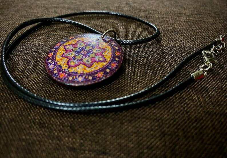 May include: A round pendant necklace with a detailed, colorful floral design in shades of purple, yellow, and orange. The pendant is attached to a black cord necklace with a silver-tone clasp.