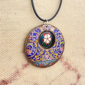 May include: A circular pendant necklace with a floral design. The pendant features a blue and gold pattern with a central flower design in red, white, and green. The necklace has a black cord.