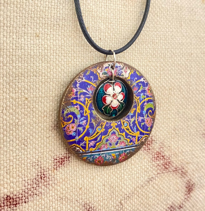May include: A circular pendant necklace with a floral design. The pendant features a blue and gold pattern with a central flower design in red, white, and green. The necklace has a black cord.