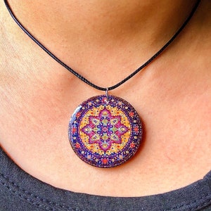 May include: A round pendant necklace with a colorful, intricate pattern resembling a Persian rug. The pendant is primarily blue, yellow, and purple, with a black cord. The necklace is worn around the neck.