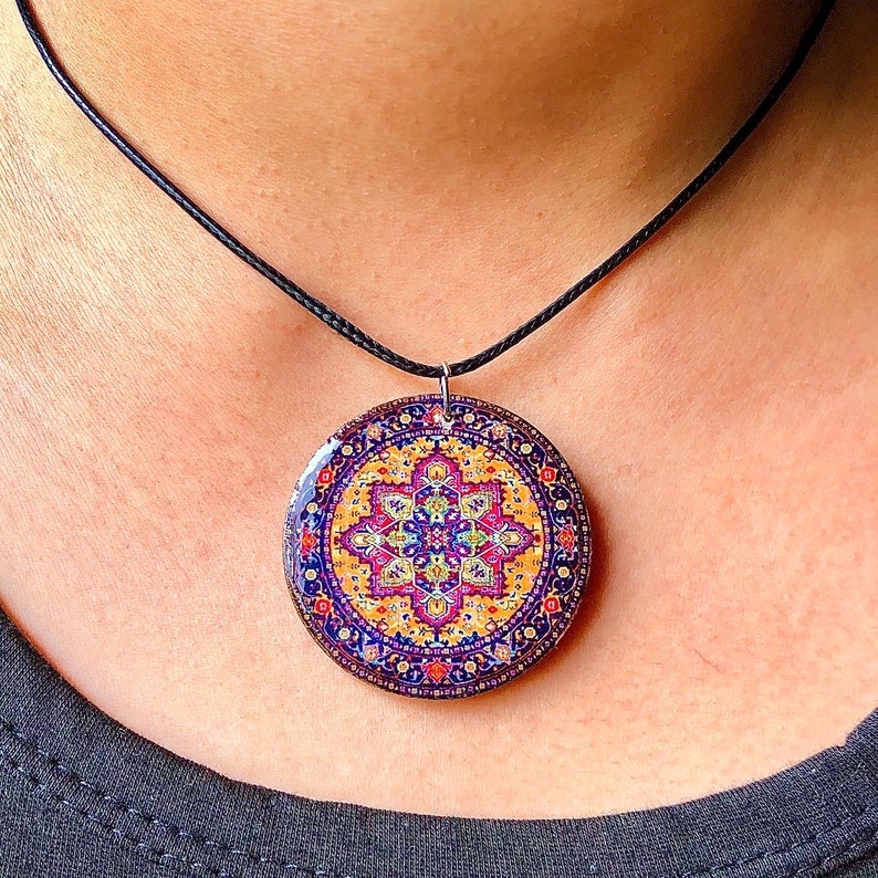 May include: A round pendant necklace with a colorful, intricate pattern resembling a Persian rug. The pendant is primarily blue, yellow, and purple, with a black cord. The necklace is worn around the neck.