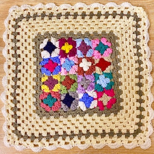 以下が含まれることがあります： 手作りの正方形のかぎ針編みブランケット。中央には、ピンク、青、赤、黄色の花柄のパッチワークが施されています。クリーム色のスカラップエッジがあり、約61cm四方です。
