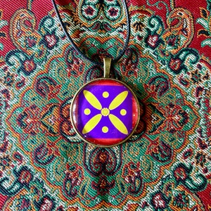 Op de afbeelding: Een ronde hangerketting met een bronzen frame. De hanger heeft een paarse achtergrond met een geel bloemmotief. De ketting zit aan een zwart koord, tegen een patroon van rode en groene stof.