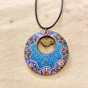 May include: A pendant necklace featuring a circular, patterned pendant with a central bird charm. The pendant has a blue, orange, and white mosaic design. The necklace has a black cord.