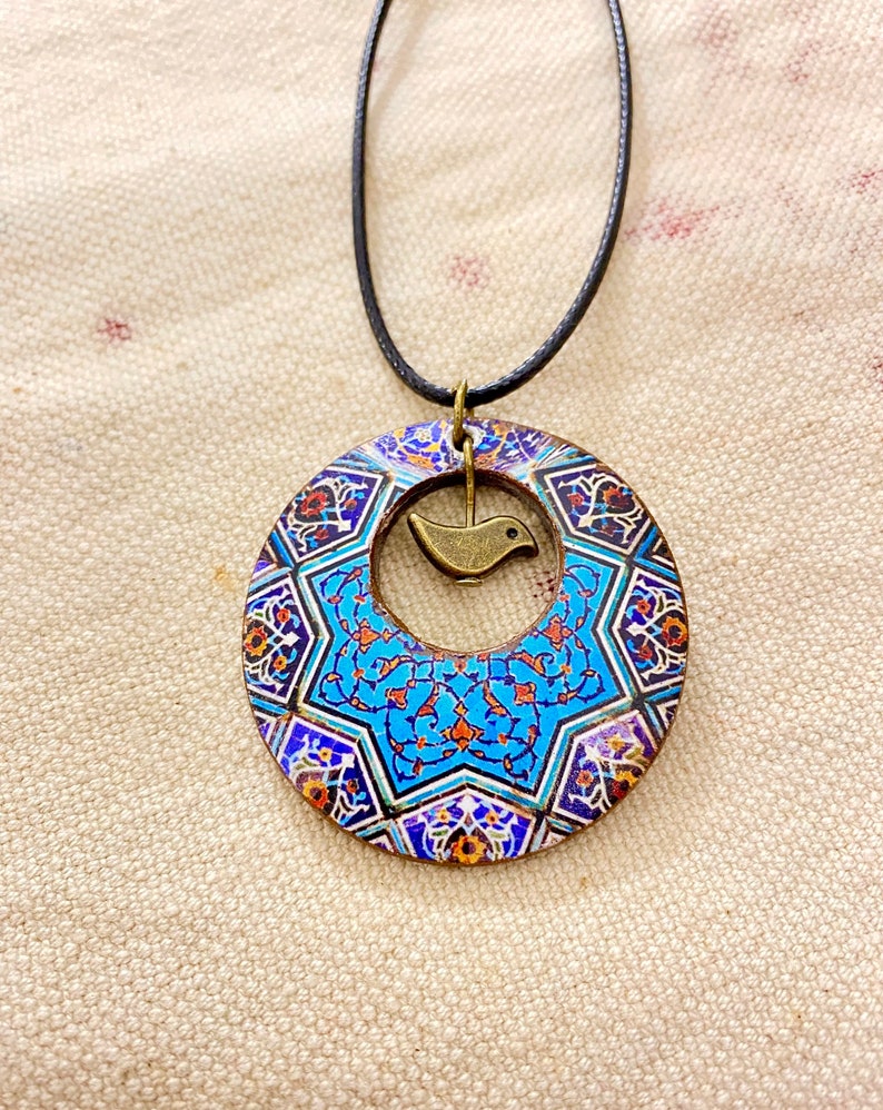 May include: A pendant necklace featuring a circular, patterned pendant with a central bird charm. The pendant has a blue, orange, and white mosaic design. The necklace has a black cord.