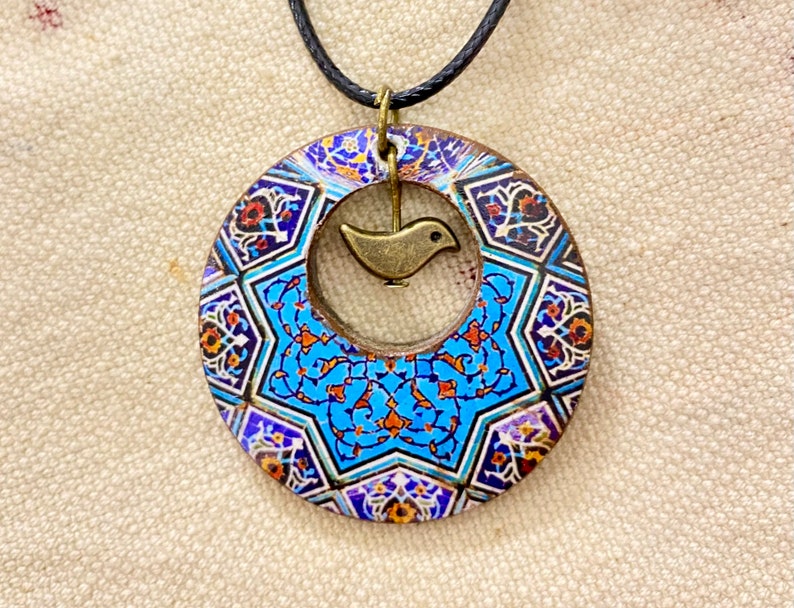 May include: A circular pendant necklace with a detailed mosaic pattern in blue, orange, and white. A small, bronze-colored bird charm hangs in the center of the pendant. The necklace has a black cord.