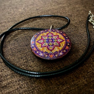 May include: A round pendant necklace with a colorful, patterned design. The pendant features a detailed, circular pattern in shades of purple, yellow, and pink. The pendant hangs from a black cord necklace with a silver clasp.