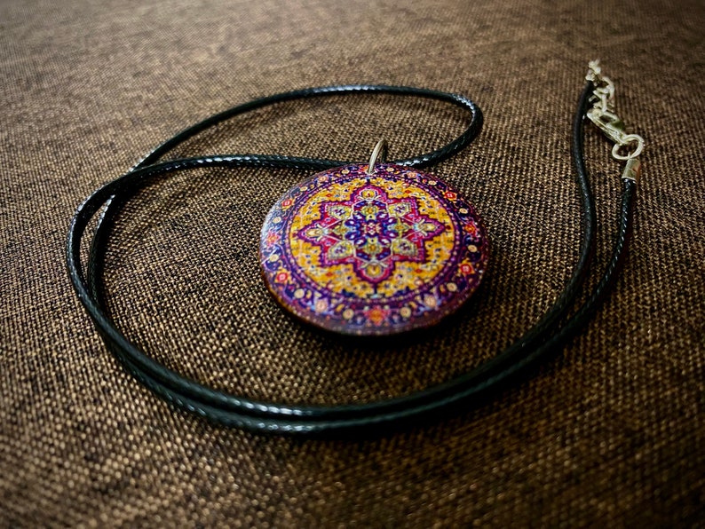 May include: A round pendant necklace with a colorful, patterned design. The pendant features a detailed, circular pattern in shades of purple, yellow, and pink. The pendant hangs from a black cord necklace with a silver clasp.