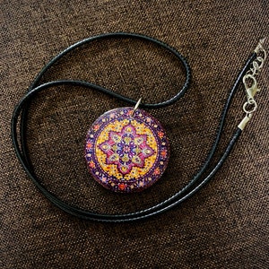 May include: A round pendant necklace with a black cord. The pendant features a colorful, intricate design with purple, yellow, and pink hues. The necklace has a silver clasp.