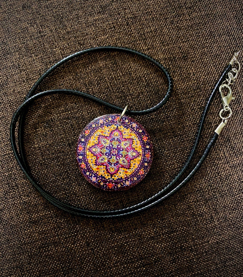 May include: A round pendant necklace with a black cord. The pendant features a colorful, intricate design with purple, yellow, and pink hues. The necklace has a silver clasp.