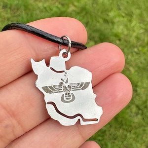 Può includere: Una collana con pendente in argento raffigurante il contorno dettagliato della mappa di un paese. Il pendente ha un cordino nero e un emblema dettagliato al centro. La collana è tenuta nella mano di una persona su uno sfondo erboso verde.