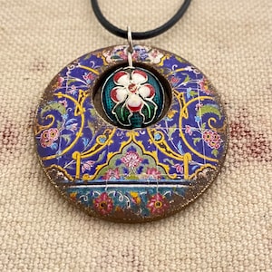 May include: A round pendant with a floral design in blue, gold, and pink. The pendant has a central opening with a smaller, round, floral-decorated element. The pendant hangs from a black cord.