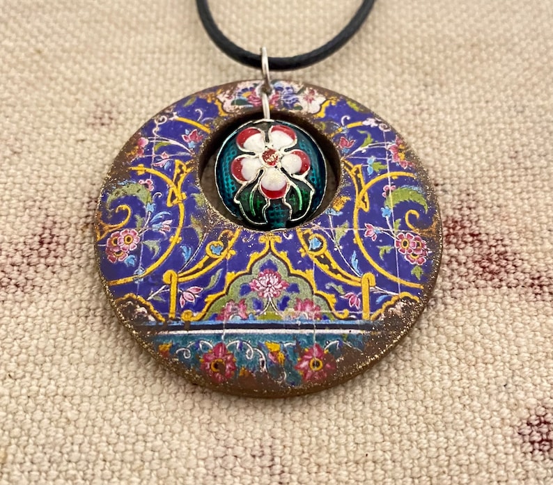 May include: A round pendant with a floral design in blue, gold, and pink. The pendant has a central opening with a smaller, round, floral-decorated element. The pendant hangs from a black cord.