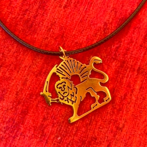 Puede incluir: Collar con colgante dorado que representa un león sosteniendo una espada, con un diseño de rayos de sol en la parte superior. El colgante está suspendido de un cordón marrón oscuro, sobre un fondo rojo.